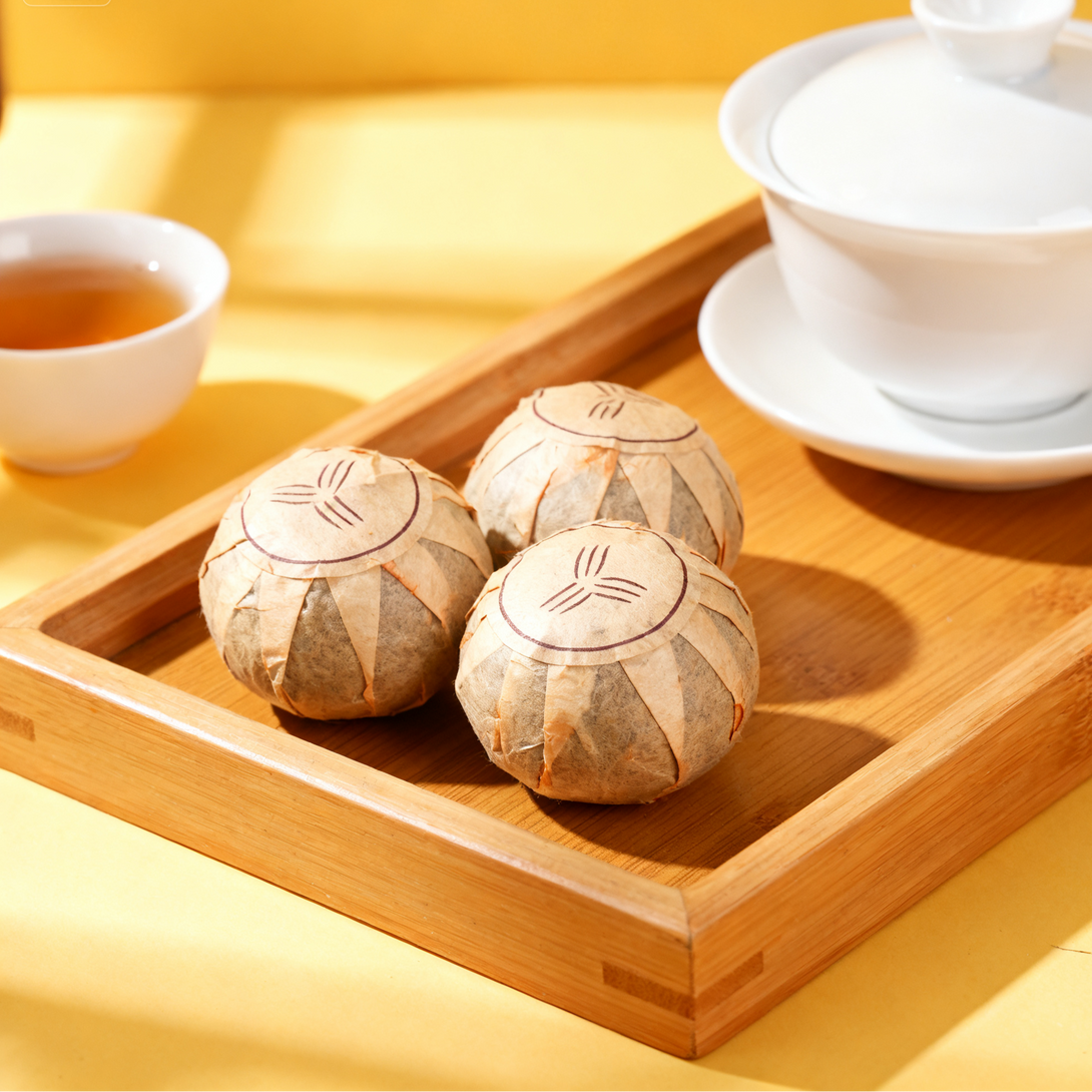 Three wrapped mini Pu-erh tea cakes on a wooden tea tray with gaiwan and teacup.