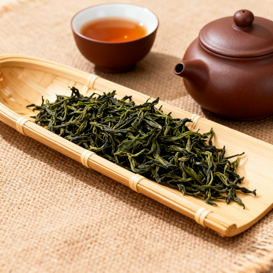 Loose green tea leaves on a bamboo tray with a teapot and cup in the background.