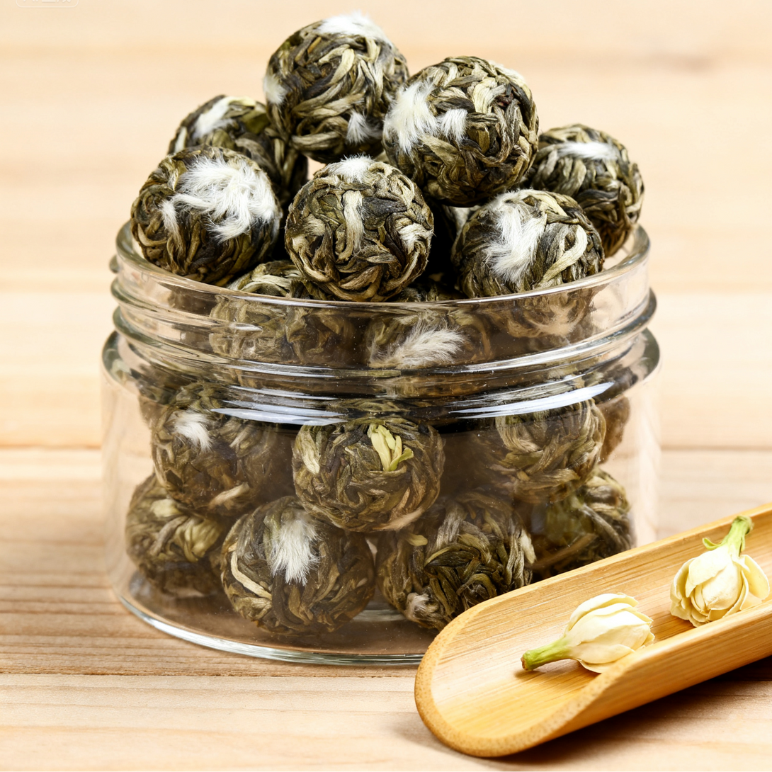 Rolled green tea balls in a clear glass jar with jasmine flowers beside it on a wooden surface