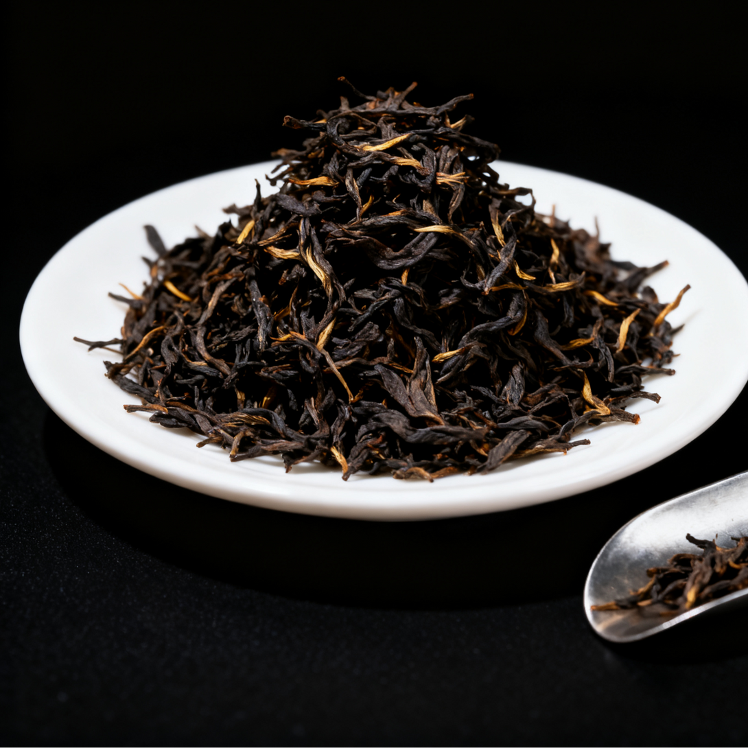A close-up view of a mound of finely crafted black tea leaves placed on a white ceramic plate, set against a dark background. The tea leaves are well-processed and dried, exhibiting a rich and glossy texture with a mix of dark and golden hues, characteristic of premium black tea. A silver tea scoop sits beside the plate, further emphasizing the high-quality nature of the tea. This image beautifully highlights the intricate details of the tea leaves and their preparation process.