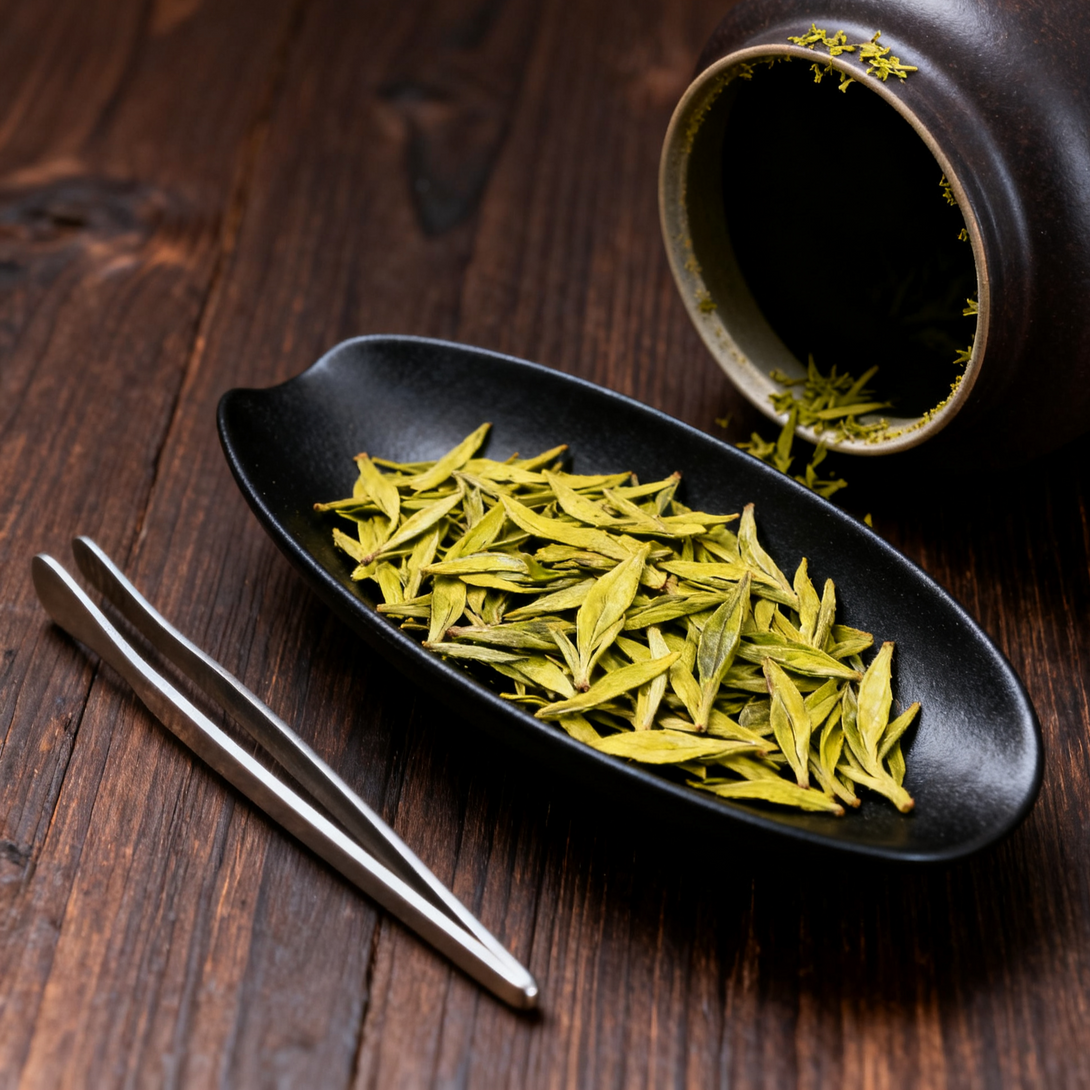 Green tea leaves on a black plate with a ceramic cup and tweezers on a wooden surface.