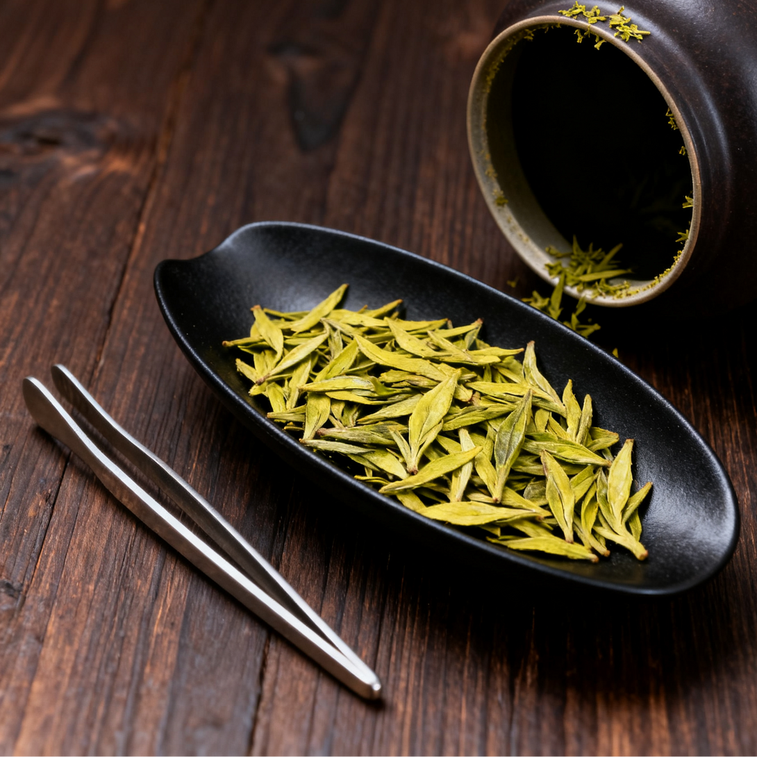 Green tea leaves arranged on a black plate with a ceramic cup and tweezers, placed on a wooden surface.
